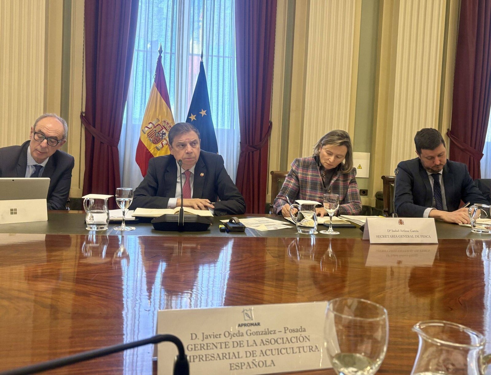 Cuatro personas sentadas en mesa de reuniones con banderas española y europea, placas identificativas y micrófonos