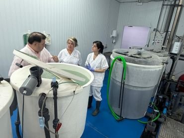 Tres personas en bata blanca junto a tanques circulares con tuberías y mangueras verdes en instalación acuicola interior