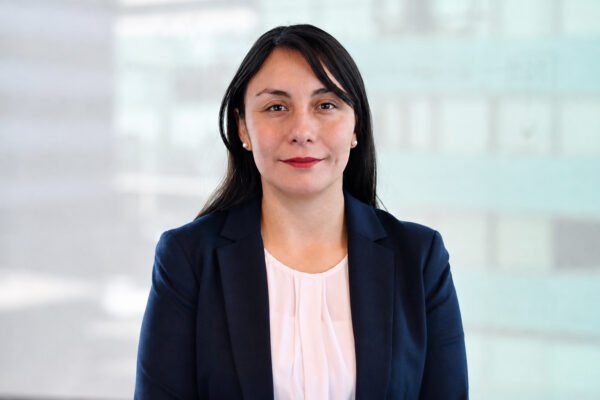 Mujer con cabello oscuro y blazer azul marino sobre blusa blanca, sonriendo suavemente frente a fondo difuminado de tonos claros