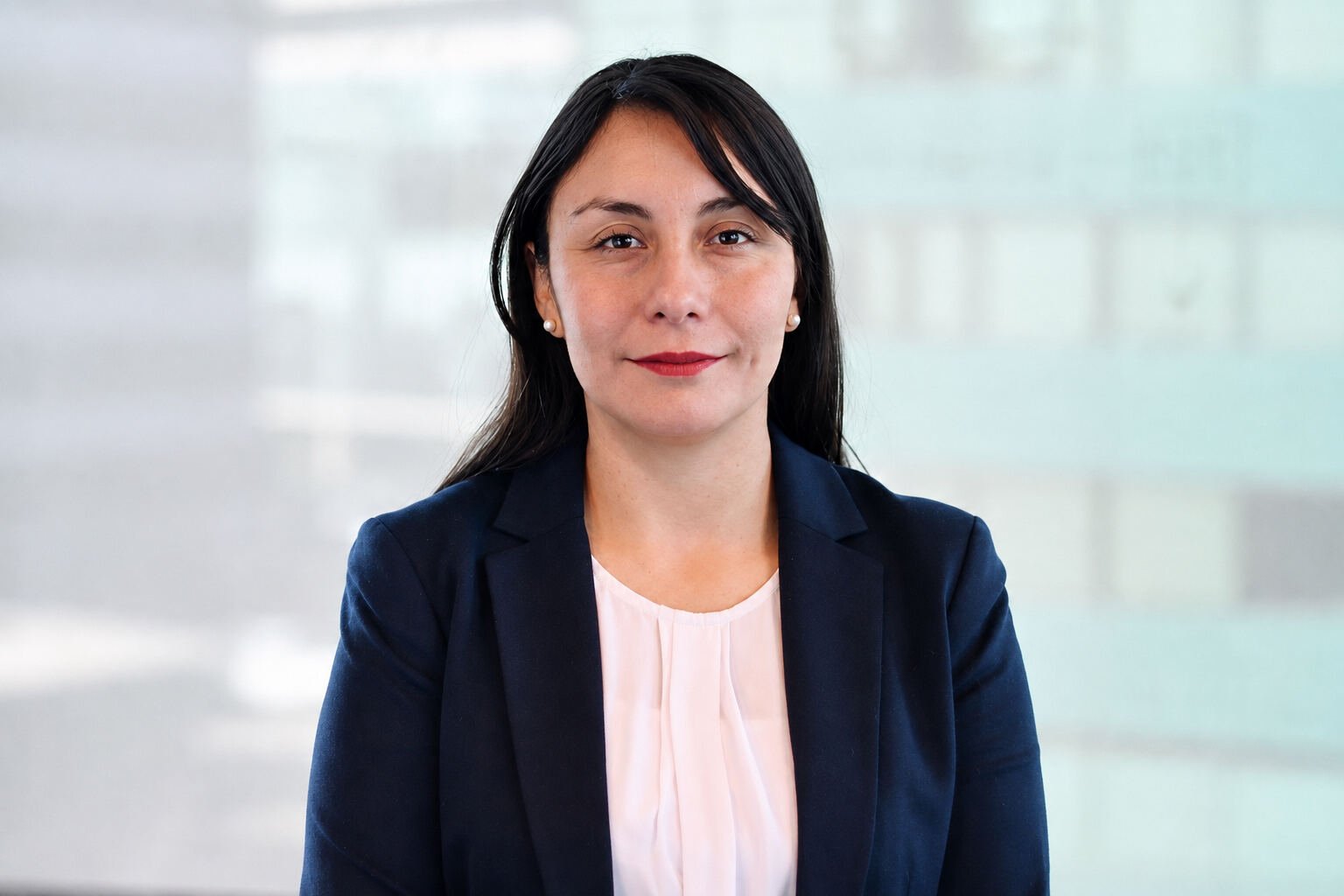 Mujer con cabello oscuro y blazer azul marino sobre blusa blanca, sonriendo suavemente frente a fondo difuminado de tonos claros